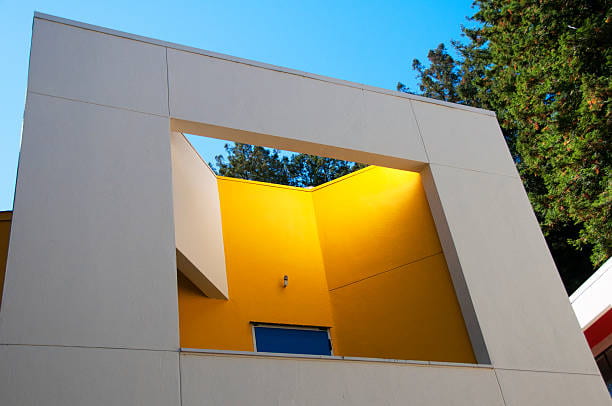 This is a photo of a dorm on the UCSC campus. It's shot from below and is a modern white square, with a center area that is gold, with a partially seen blue door. Above is a blue sky.
