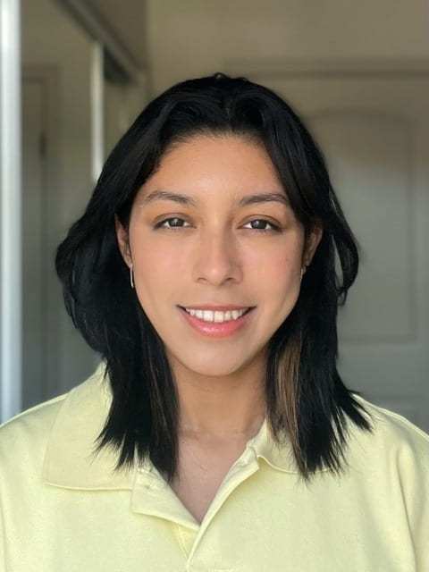 This is a portrait of Julia Spilman. They are a woman of color with dark, shoulder-length hair. They smile at the camera and are wearing a yellow polo shirt. They appear to be in some kind of hallway. 